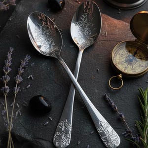 Op de afbeelding: Twee zilveren lepels met bloemmotieven kruisen elkaar op een donker oppervlak. De afbeelding bevat een brandende lantaarn, een kompas, zwarte stenen en takjes lavendel en rozemarijn, wat een mystieke esthetiek creëert.