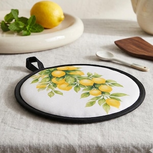 Può includere: Una presina rotonda in tessuto con sfondo bianco e un'illustrazione stampata di limoni gialli e foglie verdi su rami. La presina presenta un bordo in tessuto nero e un piccolo occhiello nero sulla parte superiore per appenderla.
