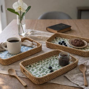 Könnte beinhalten: Drei rechteckige gewebte Tabletts mit gemusterten Einsätzen sind auf einem Holztisch angeordnet. Ein Tablett enthält eine weiße Tasse mit Kaffee, ein anderes einen Keks und das dritte ein Glas Bohnen. Holzlöffel und eine Blumenvase sind ebenfalls vorhanden.