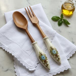 Peut inclure: Cuillère et fourchette en bois avec des manches crème ornés de motifs floraux bleus et verts. Les ustensiles reposent sur une serviette en lin blanche à bordure en dentelle. Une petite bouteille d'huile et des feuilles de basilic sont en arrière-plan.