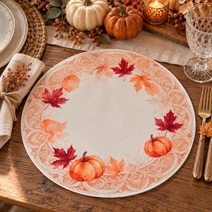 May include: A round placemat featuring a cream-colored center surrounded by a decorative border of orange and red watercolor maple leaves and pumpkins. The intricate design includes swirling scrollwork patterns in soft peach and light orange tones.