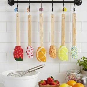 Può includere: Un set di sette spatole in legno con teste in silicone a tema frutta, tra cui fragole, arance e limoni, appese a una barra di metallo nero. Una ciotola bianca, una frusta e frutta fresca in primo piano.