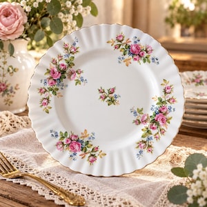 Peut inclure: Une assiette en céramique blanche avec un bord festonné et une bordure dorée. L'assiette présente un motif floral composé de roses roses, de feuilles vertes et de petites fleurs bleues disposées en grappes autour du centre et sur le bord cannelé.