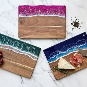 Peut inclure: Trois planches à découper en bois avec des motifs de vagues en résine bleues, vertes et violettes. Chaque planche a une base en bois naturel et un dessus en résine imitant les vagues de l'océan. Une planche a du fromage et de la viande.