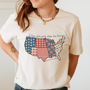 May include: Cream-colored t-shirt featuring a patchwork design of the United States map. The map incorporates various patterns, including stars, floral prints, and gingham. The shirt has the text "To Know God and to Make Him Known" and "Classical conversations".