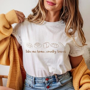 May include: White t-shirt with a brown illustration of various bread loaves and the text "Take me home, country loaves." The shirt is being worn with a yellow cardigan and blue jeans.