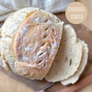 May include: A loaf of sourdough bread with decorative leaf-shaped cuts, partially sliced, on a wooden cutting board. The bread has a light crust and is dusted with flour. A circular label reads "SOURDOUGH STARTER".