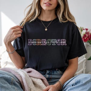 May include: Black t-shirt with the word "DIRECTOR" in a colorful, outlined font. Below the word "DIRECTOR" is the text "Classical Conversations". The shirt is a crew neck style.