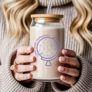 May include: A clear glass tumbler with a bamboo lid and a glass straw, filled with a creamy beverage. The tumbler features a purple globe design with the words "Home Sweet Homeschool" and floral accents. The tumbler is held by a person.