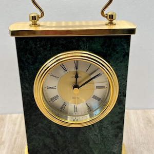 May include: A rectangular mantel clock featuring a dark green marble-effect body with gold-tone metal accents. The clock face is circular with a gold center, black Roman numerals, and black hands. It includes a gold handle and base.