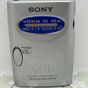 May include: A silver Sony Walkman SRF-59 FM/AM radio with a blue oval display showing frequency scales for FM and AM bands. The device features a power switch with on and off labels, a tuning indicator, and the Walkman logo printed on the front.