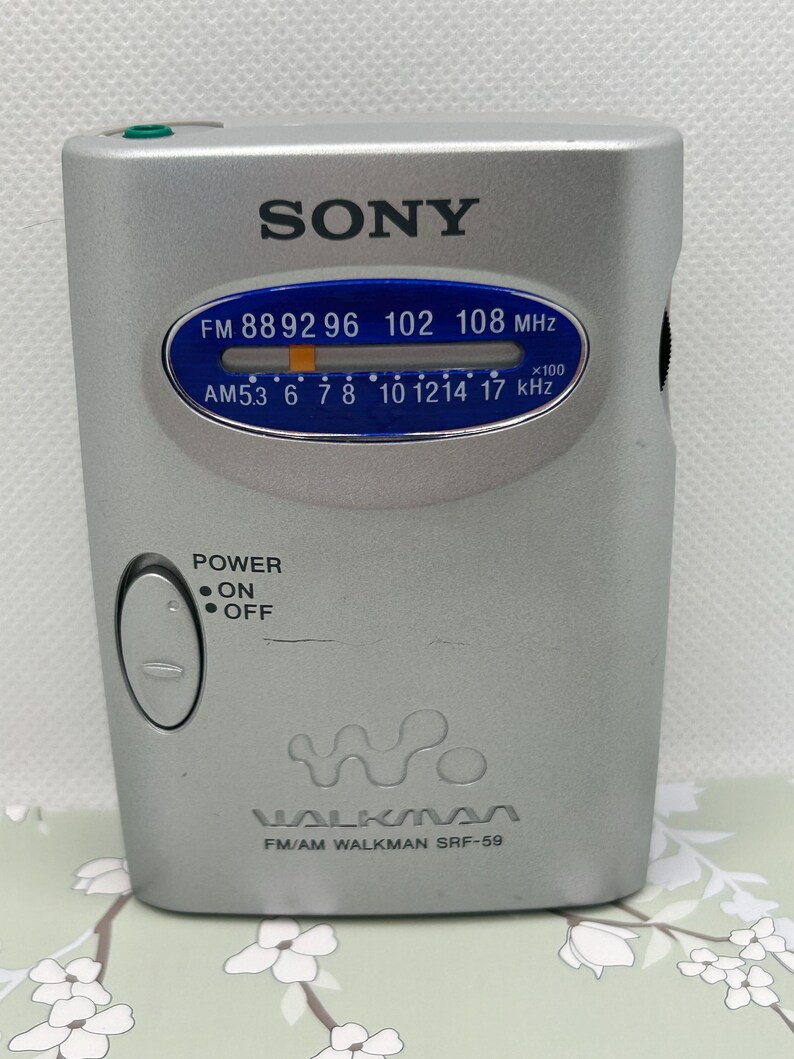 May include: A silver Sony Walkman SRF-59 FM/AM radio with a blue oval display showing frequency scales for FM and AM bands. The device features a power switch with on and off labels, a tuning indicator, and the Walkman logo printed on the front.