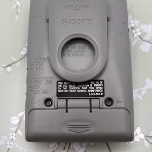 May include: A gray Sony portable radio featuring a belt clip, a battery compartment, and controls labeled TUNE, VOL, BAND FM AM, and FM SENS DX LOCAL. The back includes text regarding FCC compliance and battery requirements for 1.5V R6 size AA batteries.