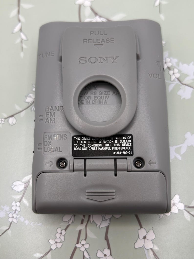 May include: A gray Sony portable radio featuring a belt clip, a battery compartment, and controls labeled TUNE, VOL, BAND FM AM, and FM SENS DX LOCAL. The back includes text regarding FCC compliance and battery requirements for 1.5V R6 size AA batteries.