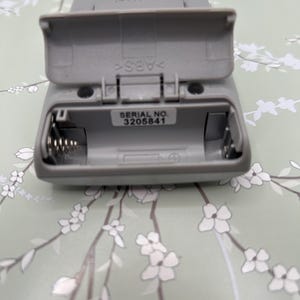 May include: A gray plastic Sony electronic device with an open battery compartment door. The interior reveals a metal spring contact and a white label with the text SERIAL NO. 3205841. The plastic casing is embossed with the word SONY and ABS.