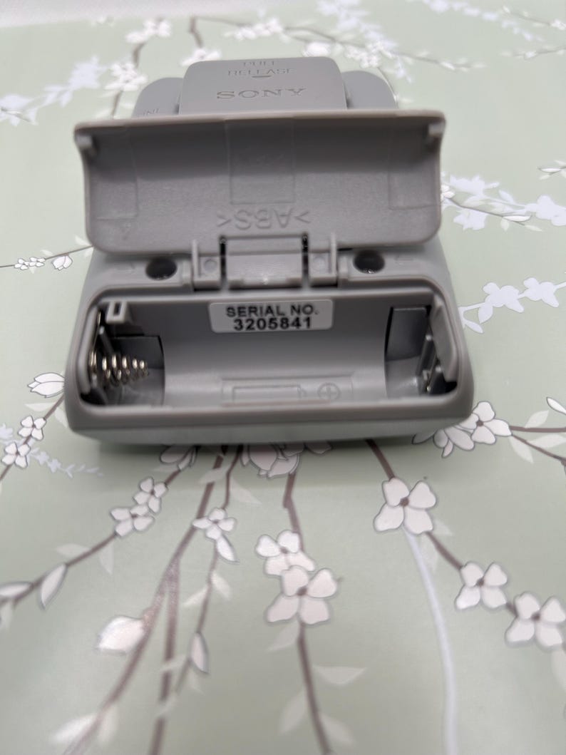May include: A gray plastic Sony electronic device with an open battery compartment door. The interior reveals a metal spring contact and a white label with the text SERIAL NO. 3205841. The plastic casing is embossed with the word SONY and ABS.