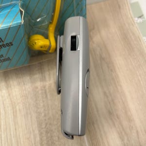 May include: A gray portable electronic device with a side-mounted black volume wheel and a clip, positioned next to a blue and yellow retail package that features partial text including the word reos and the word tant in a gold font.