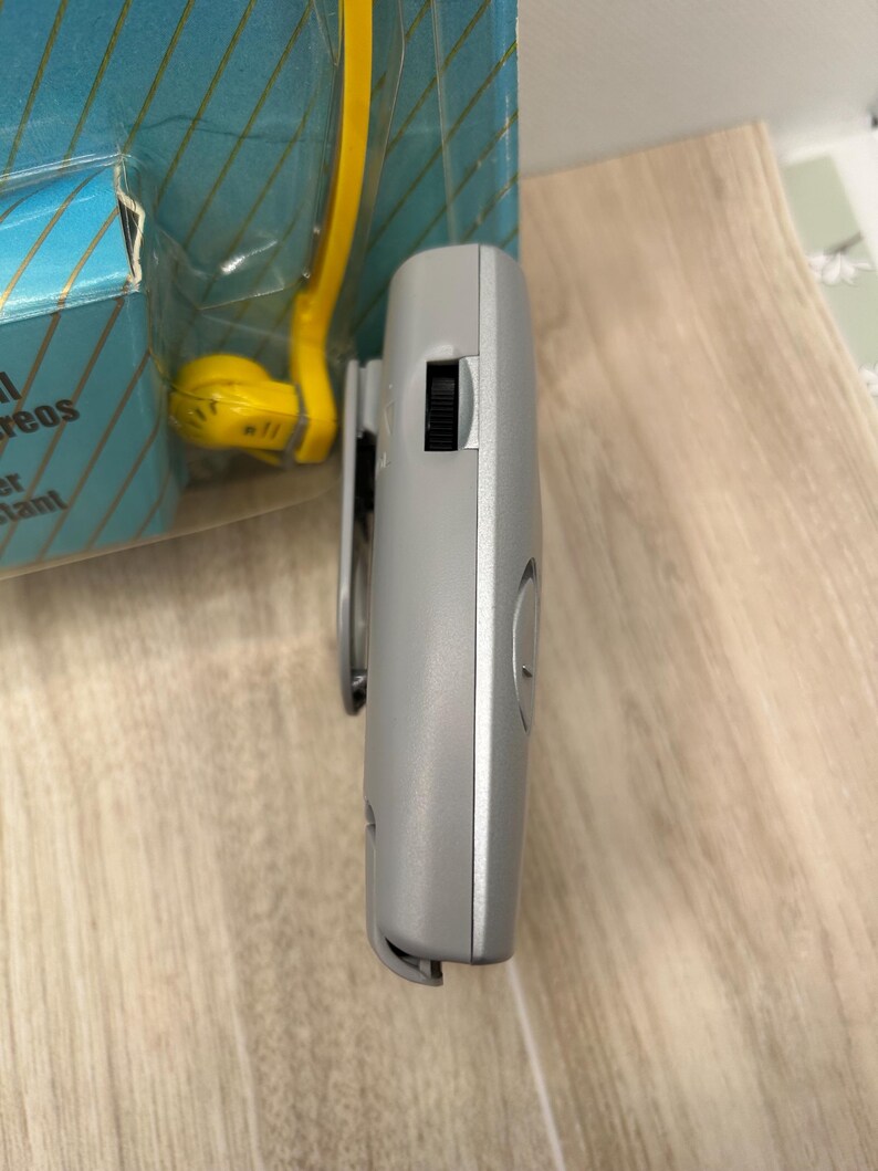 May include: A gray portable electronic device with a side-mounted black volume wheel and a clip, positioned next to a blue and yellow retail package that features partial text including the word reos and the word tant in a gold font.