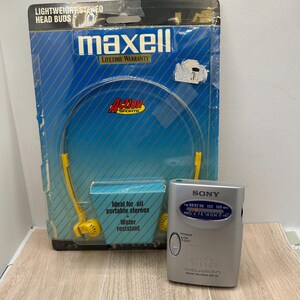 May include: A silver Sony Walkman FM/AM radio with a power switch and frequency dial, positioned next to a blue Maxell Action Sports packaging containing yellow lightweight stereo head buds with a water resistant label.