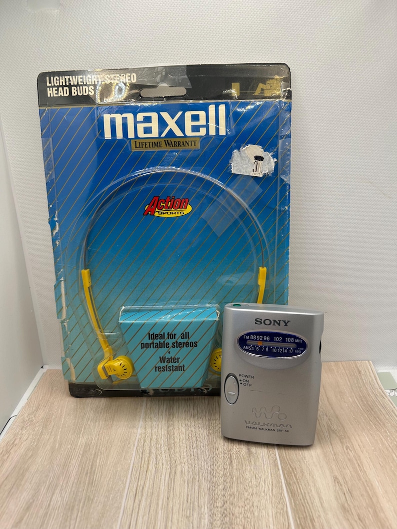 May include: A silver Sony Walkman FM/AM radio with a power switch and frequency dial, positioned next to a blue Maxell Action Sports packaging containing yellow lightweight stereo head buds with a water resistant label.