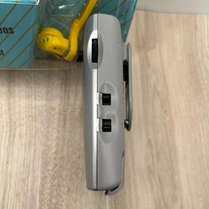 May include: A silver portable electronic device with a side-mounted belt clip, a black volume dial, and two black sliding switches. The device is positioned vertically in front of its original blue and yellow retail packaging.