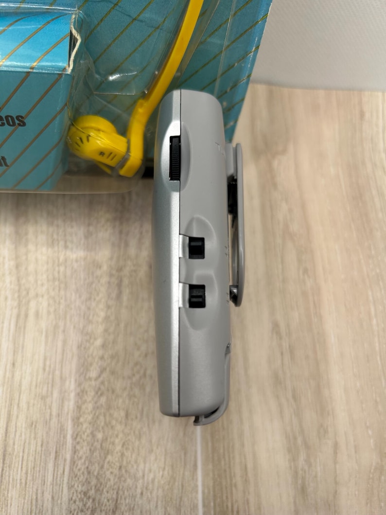 May include: A silver portable electronic device with a side-mounted belt clip, a black volume dial, and two black sliding switches. The device is positioned vertically in front of its original blue and yellow retail packaging.