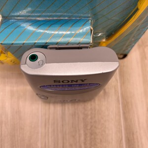 May include: A silver Sony portable FM radio with a top-mounted green headphone jack and a blue frequency display panel showing numbers from 88 to 108. The device is partially encased in its original blue and yellow cardboard packaging.