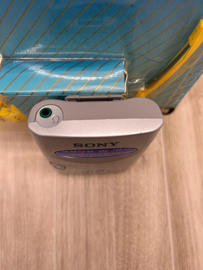 May include: A silver Sony portable FM radio with a top-mounted green headphone jack and a blue frequency display panel showing numbers from 88 to 108. The device is partially encased in its original blue and yellow cardboard packaging.