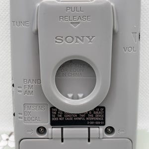 May include: A light gray Sony portable radio featuring a belt clip with PULL RELEASE text. The back panel displays TUNE, VOL, BAND FM AM, and FM SENS DX LOCAL controls. It includes battery compartment details and FCC compliance text printed on the casing.