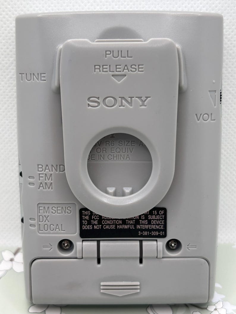 May include: A light gray Sony portable radio featuring a belt clip with PULL RELEASE text. The back panel displays TUNE, VOL, BAND FM AM, and FM SENS DX LOCAL controls. It includes battery compartment details and FCC compliance text printed on the casing.