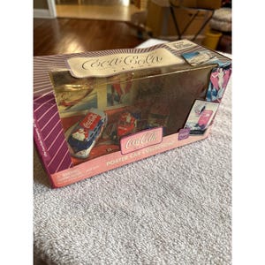 May include: A Coca-Cola Poster Car Collection box set featuring a clear plastic window displaying miniature die-cast vehicles. The packaging is purple and white with vintage-style graphic advertisements, including a Coca-Cola logo and text.