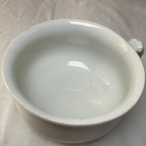 May include: A white ceramic chamber pot with a smooth, rounded basin and a single small handle on the side. The vessel features a wide, open top and a deep, curved interior, with subtle signs of wear along the upper rim and base edges.