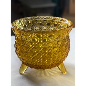 May include: A small, amber-colored glass bowl featuring a textured, pressed pattern of raised circles and cross-hatched lines. The bowl is supported by three short, smooth glass feet and has a wide, circular opening with a slightly flared rim.