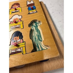 May include: Wooden puzzle featuring a school theme. The puzzle has cutouts of students at desks and a tree. The students are depicted with colorful clothing and facial features. The puzzle is made of light brown wood.
