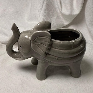 May include: A gray ceramic elephant planter with a large opening on its back. The elephant has detailed features, including large ears, a curled trunk, and small legs. The planter is designed to hold plants or other decorative items.