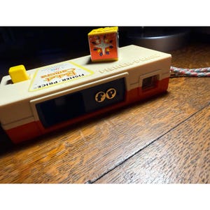 May include: A vintage Fisher-Price Pocket Camera toy in cream, red, and yellow. The toy camera has a black lens area, the Fisher-Price logo, and a small yellow cube on top. A colorful cube with a starburst design sits on top.