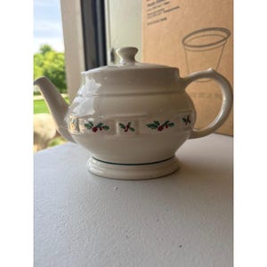 May include: Cream-colored ceramic teapot with a lid and handle. The teapot features a decorative band with holly and berry designs. A green line accents the base. The teapot is designed for brewing tea.