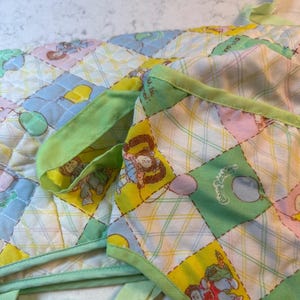 May include: Close-up of a quilted baby blanket with a patchwork design. The squares feature cartoon characters, balloons, and pastel colors like blue, yellow, and pink. The edges are trimmed with a light green fabric.