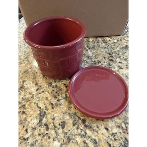 May include: A deep red ceramic crock with a cylindrical shape and a matching circular lid. The exterior of the crock features a subtle embossed brick-like pattern around the middle section, and the entire surface has a smooth, glossy finish.