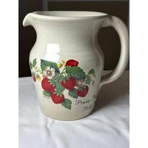 May include: A cream-colored ceramic pitcher featuring a botanical illustration of red strawberries, green leaves, and white blossoms. The text "Praise His Holy Name" is printed in black script near the base, along with the artist signature D. Hagal.
