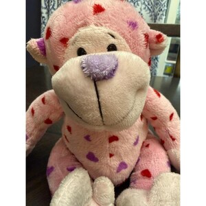May include: A plush pink monkey with red and purple heart-shaped accents. The monkey has a light purple nose and ears, and black eyes. The toy is soft and cuddly, perfect for children.