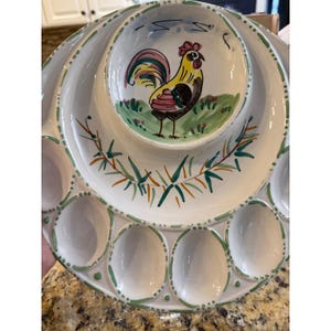 May include: A white ceramic serving platter featuring a central bowl painted with a yellow, pink, and brown rooster standing on green grass. The platter has a circular arrangement of twelve egg-shaped indentations rimmed with green painted dots.
