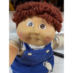 May include: A vintage Cabbage Patch Kid doll with a fair complexion, rosy cheeks, and brown yarn hair. The doll is wearing a blue denim overall and a cream-colored shirt. An embroidered elephant patch is on the overall.