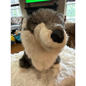 May include: A plush wolf toy featuring soft, shaggy fur in shades of gray and white. The stuffed animal has a prominent, smooth black nose, rounded ears, and a thick, textured coat that covers its head and chest area in a realistic pattern.
