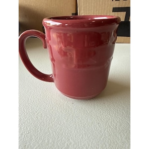 May include: A burgundy ceramic mug with a curved handle. The mug has a slightly flared rim and a smooth, glossy finish. The mug is sitting on a white surface.