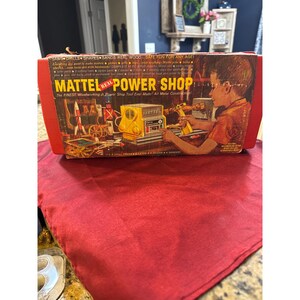 May include: A vintage Mattel Real Power Shop toy set in a red box. The box features an illustration of a child using the yellow power tool, with text reading Mattel Real Power Shop, The Finest Woodworking Jr. Power Shop Tool Ever Made.