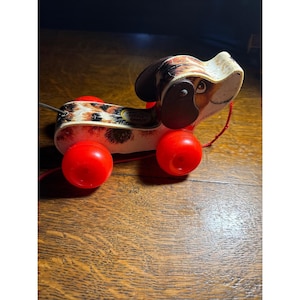 May include: Vintage Fisher-Price Little Snoopy pull toy. The toy dog has a spotted pattern on its body and head, with red wheels and a red pull string. The dog's ears and eyes are brown. The toy is on a wooden surface.