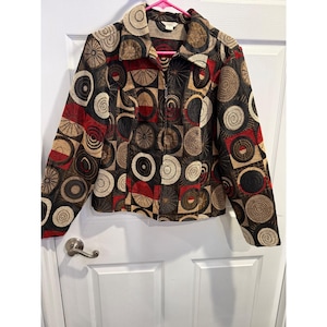May include: A zip-up jacket with a unique geometric pattern in red, black, tan, and brown. The jacket features a collar and long sleeves. The design includes circles and squares, creating a bold, eye-catching look. The jacket is hanging on a pink hanger.