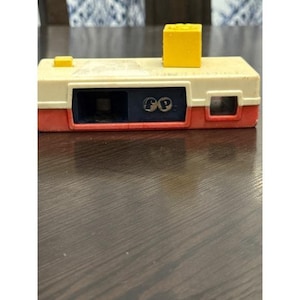 May include: A vintage toy camera in a rectangular shape with a cream-colored body, red base, and a yellow cube on top. The camera features a black lens area and the initials "FP".