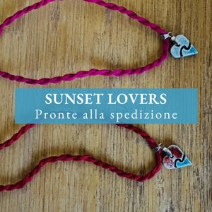 Può includere: Due collane con un cordoncino magenta intrecciato e un ciondolo a forma di cuore. Il ciondolo è un design a cuore spezzato con accenti rossi, bianchi e blu. L'immagine ha il testo "SUNSET LOVERS".